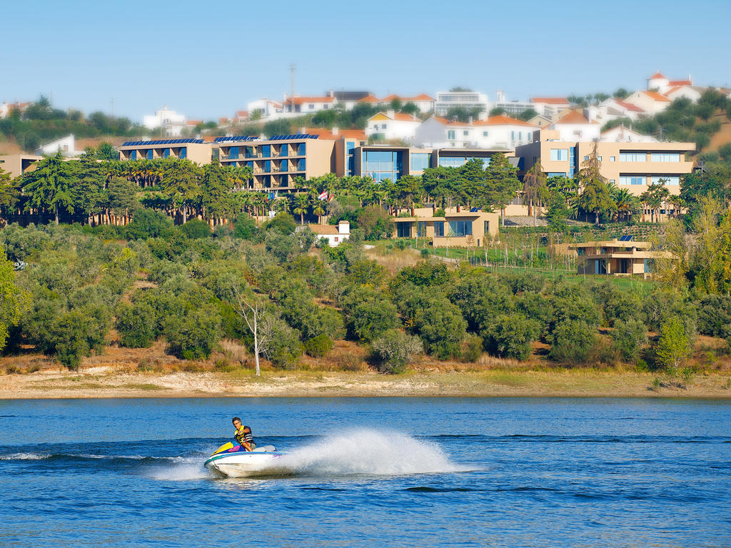 Ap Lago Montargil - Conference & Spa Exterior photo The photo depicts a person riding a jet ski on a body of water. In the background, there are modern buildings and lush greenery, suggesting a scenic landscape, likely near a resort or upscale residential area. The clear blue sky and calm water enhanc