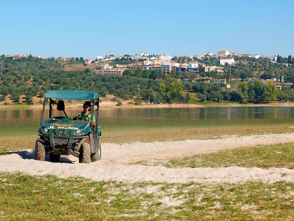 Ap Lago Montargil - Conference & Spa Exterior photo The photo shows a small green all-terrain vehicle driving along a sandy path beside a body of water. In the background, there's a landscape with trees and rolling hills, along with a town or village visible on the hillside. The sky is clear, indicati