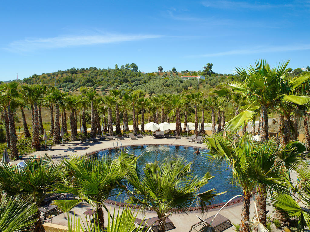 Ap Lago Montargil - Conference & Spa Exterior photo The photo depicts a beautiful outdoor area featuring a circular swimming pool surrounded by palm trees. The pool area has several lounge chairs arranged around it. In the background, there is a lush green hillside, and the sky is clear with a bright 