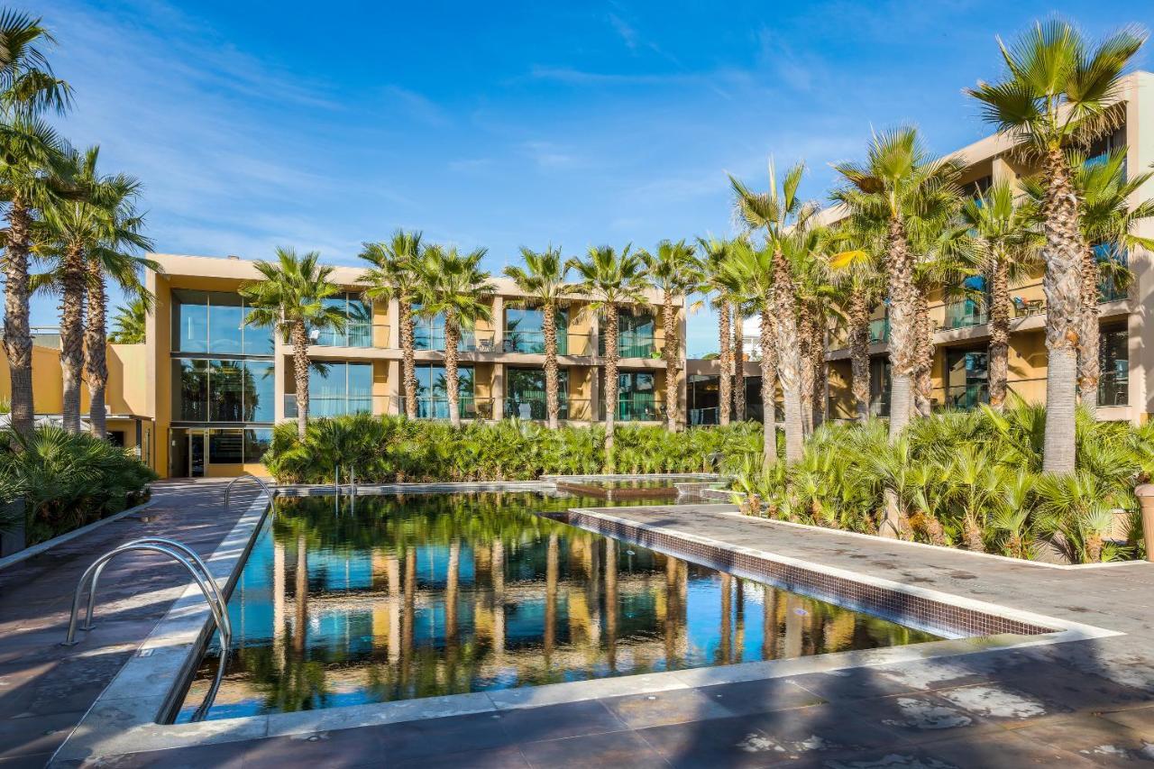 Ap Lago Montargil - Conference & Spa Exterior photo The photo features a modern building surrounded by lush greenery and palm trees. In the foreground, there is a serene swimming pool with a reflective surface that captures the surrounding environment. The architecture of the building appears to have 