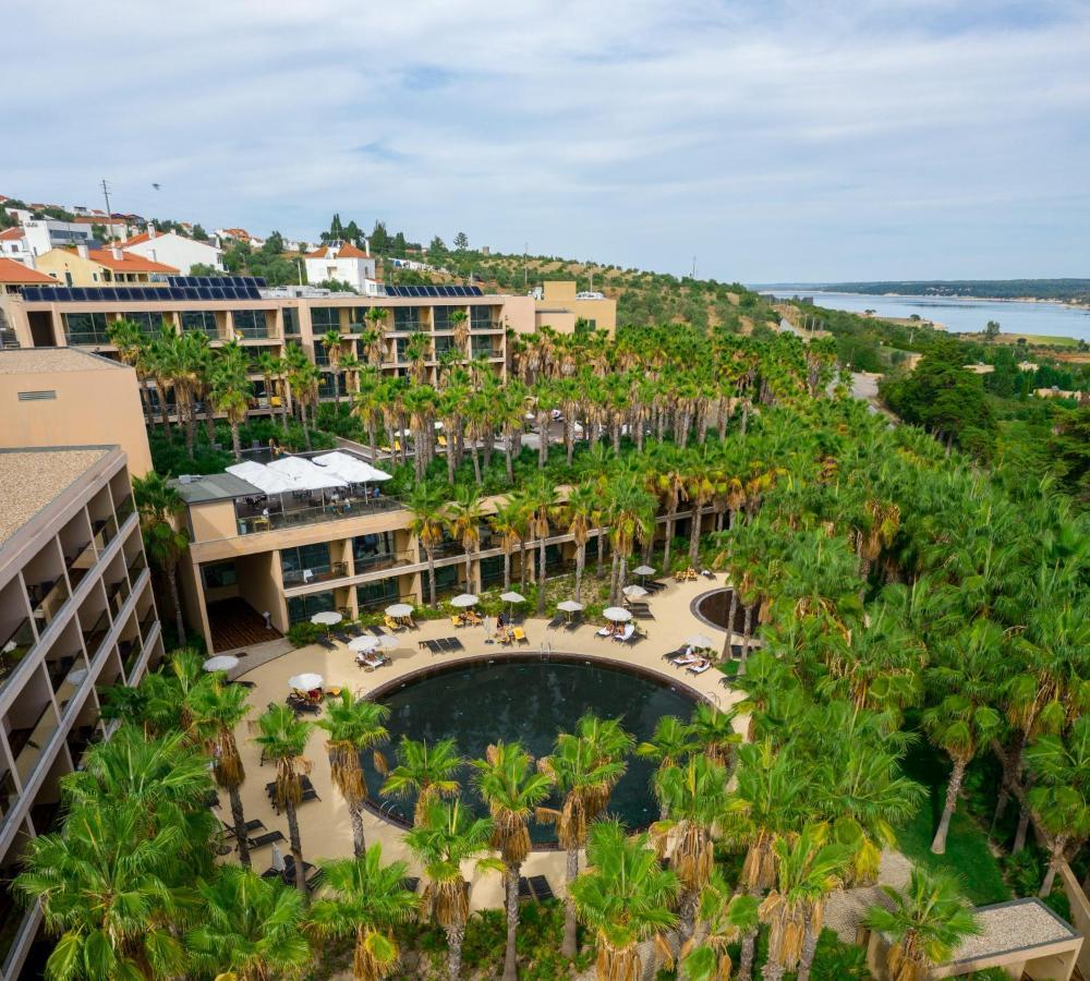 Ap Lago Montargil - Conference & Spa Exterior photo The photo shows a modern hotel complex set amidst lush greenery, featuring numerous palm trees. In the foreground, there is a circular swimming pool surrounded by lounge chairs. The hotel itself has multiple levels with balconies overlooking the pool