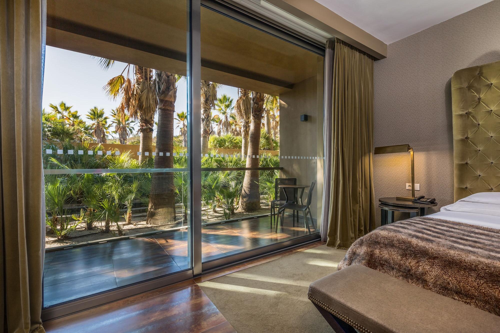 Ap Lago Montargil - Conference & Spa Exterior photo The photo shows a bedroom with large glass doors that lead out to a balcony or terrace. The view outside features palm trees and lush greenery, indicating a tropical or warm climate. Inside the room, there is a bed, a bedside table with a lamp, and a