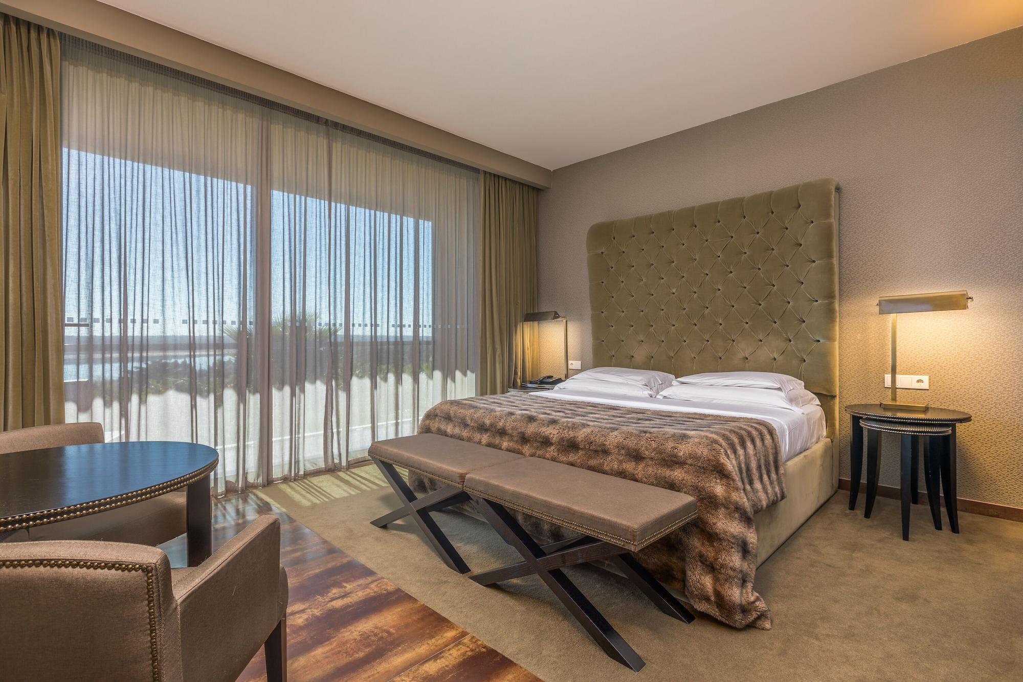 Ap Lago Montargil - Conference & Spa Exterior photo The photo shows a modern hotel room featuring a large bed with a tufted headboard and a cozy blanket. There are curtains draped over large windows that allow natural light to flow in, providing a view of the outside. A small seating area includes two