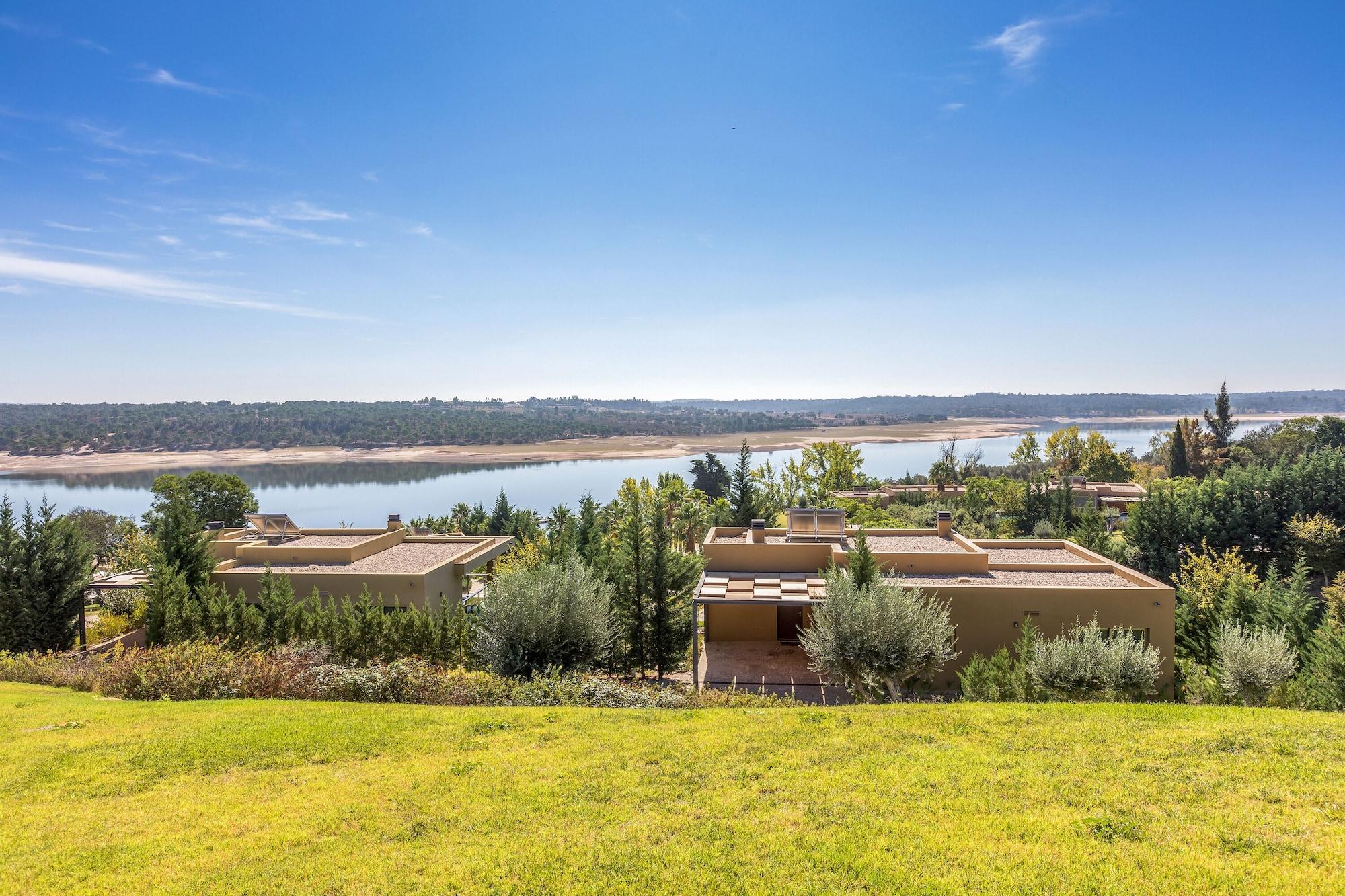 Ap Lago Montargil - Conference & Spa Exterior photo The photo depicts a scenic landscape featuring a calm body of water, possibly a lake or river, surrounded by lush greenery and trees. In the foreground, there are several modern buildings with flat roofs, set against a vibrant blue sky. The overall s