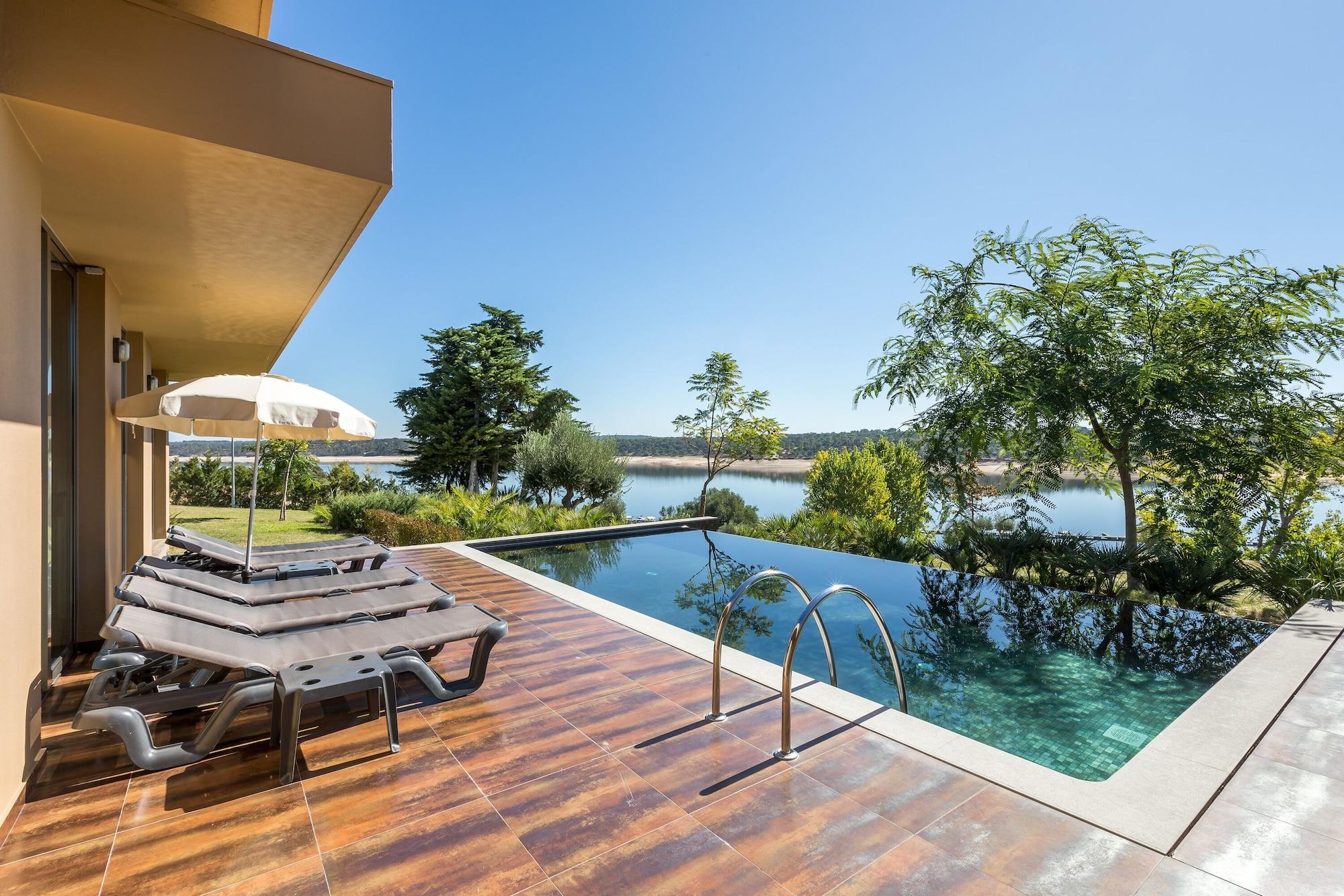 Ap Lago Montargil - Conference & Spa Exterior photo The photo shows a luxurious outdoor setting by a pool. There are several lounge chairs arranged next to an infinity pool, which overlooks a scenic body of water and lush greenery. A parasol provides shade for relaxation. The sky is clear and blue, in