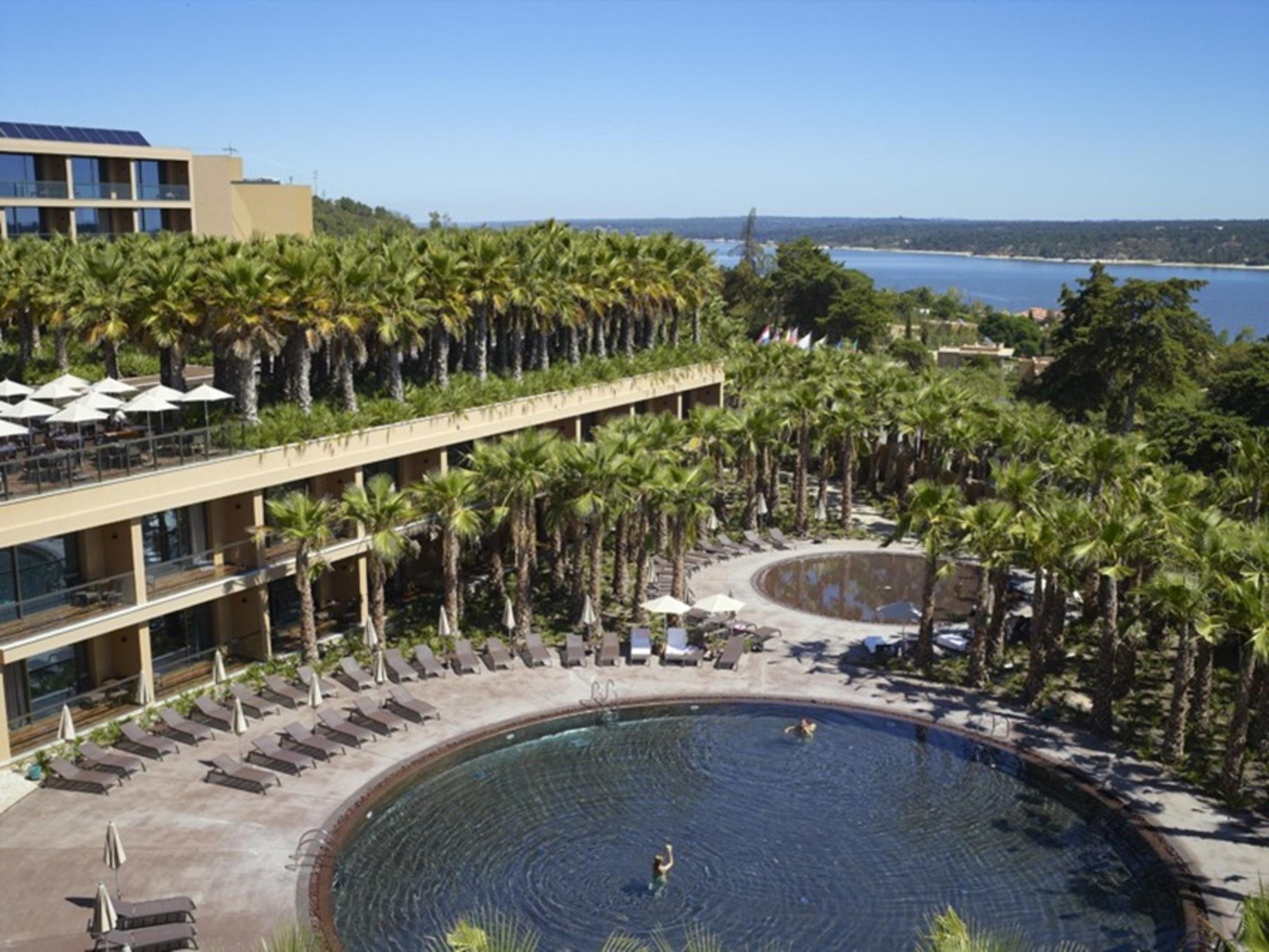 Ap Lago Montargil - Conference & Spa Exterior photo The photo shows a scenic view of a resort area. In the foreground, there are circular pools surrounded by lounge chairs, with a few people enjoying the water. Palm trees line the edges, creating a tropical atmosphere. In the background, there is a mu