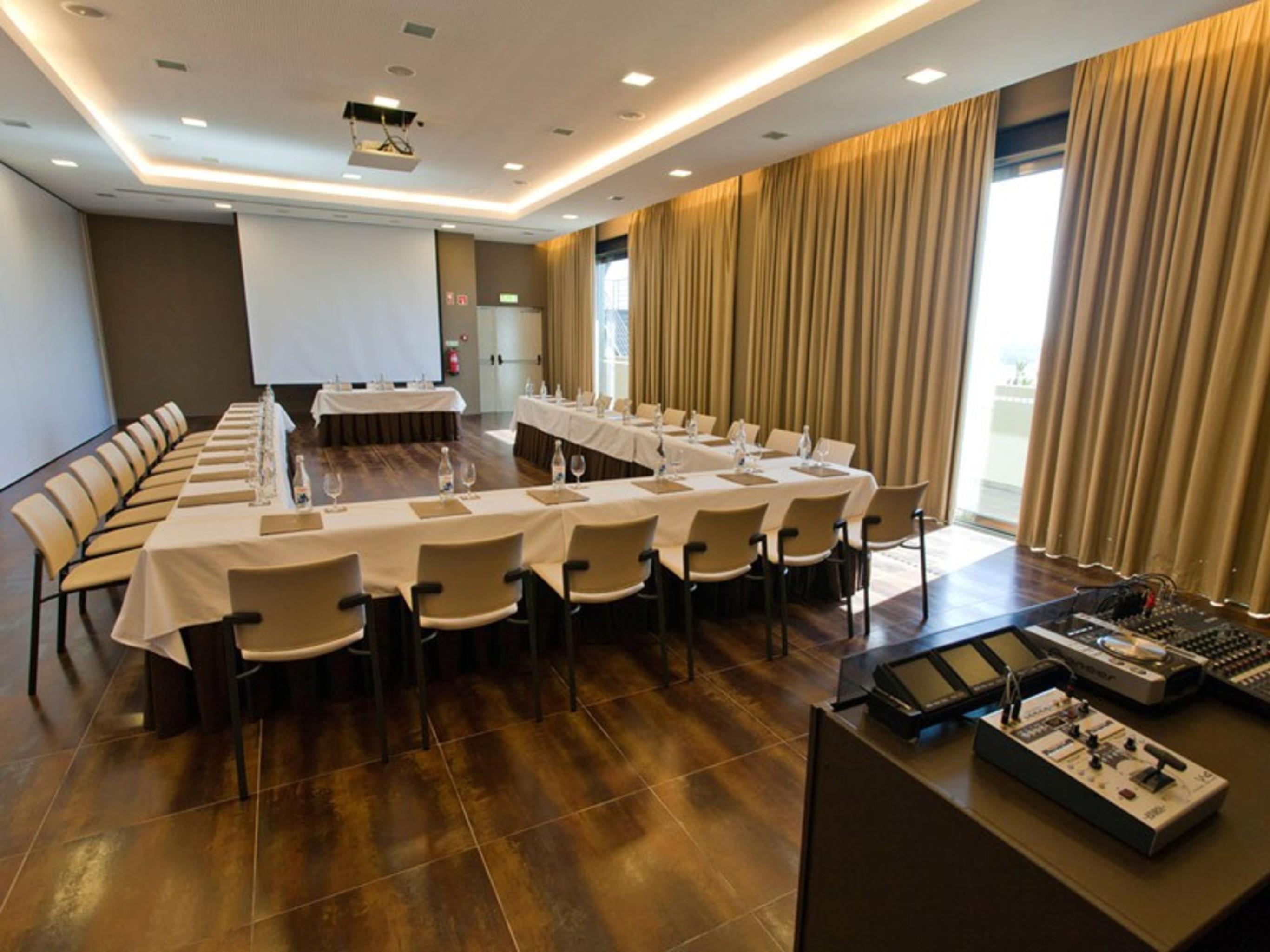 Ap Lago Montargil - Conference & Spa Exterior photo The photo shows a modern conference room set up for a meeting. There are several tables arranged in a U-shape, surrounded by chairs. A large screen and projector are positioned at the front, with a table for speakers or presenters. The room has large