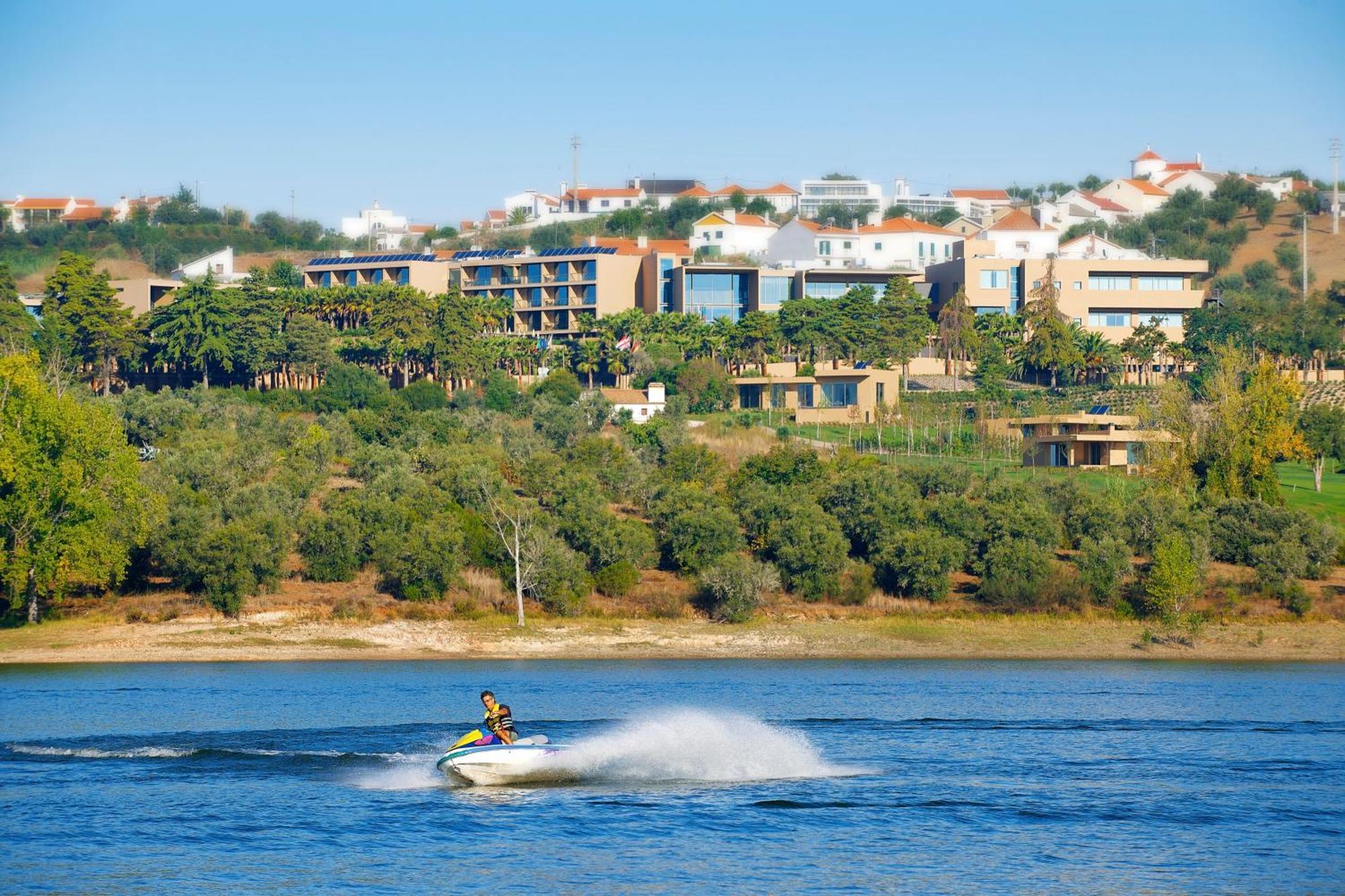 Ap Lago Montargil - Conference & Spa Exterior photo The photo shows a person riding a jet ski on a body of water, creating a spray as they speed across the surface. In the background, there are modern buildings and a hillside with trees and greenery. The sky is clear and blue, suggesting a bright and 