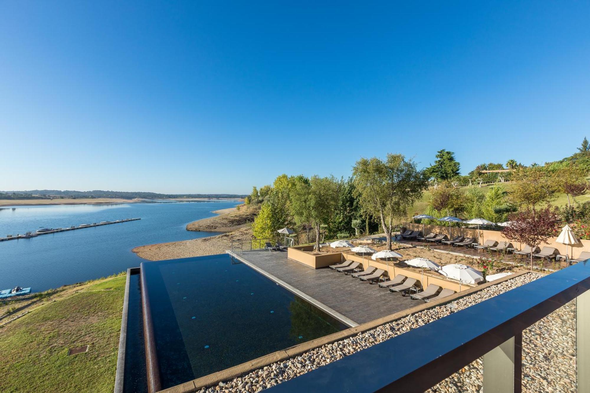 Ap Lago Montargil - Conference & Spa Exterior photo The photograph depicts a scenic view of a tranquil water body, likely a lake or river, bordered by lush greenery. In the foreground, there is an infinity pool that blends seamlessly with the water beyond. Comfortable lounge chairs are arranged on a w