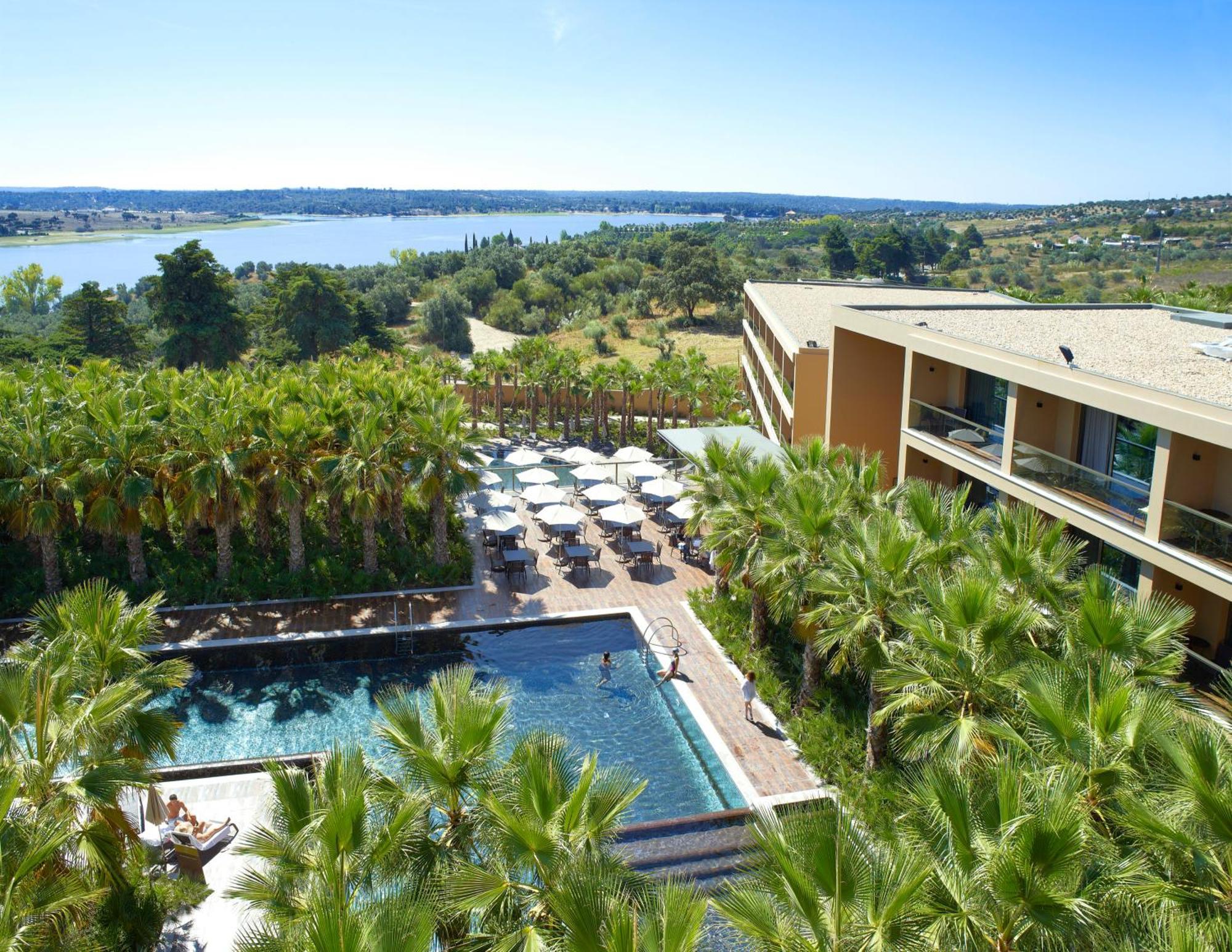 Ap Lago Montargil - Conference & Spa Exterior photo The photo shows a scenic view of a modern hotel or resort. In the foreground, there is a swimming pool surrounded by palm trees, with lounge chairs positioned beside it. Sun umbrellas provide shade over the chairs. The background features a lush land