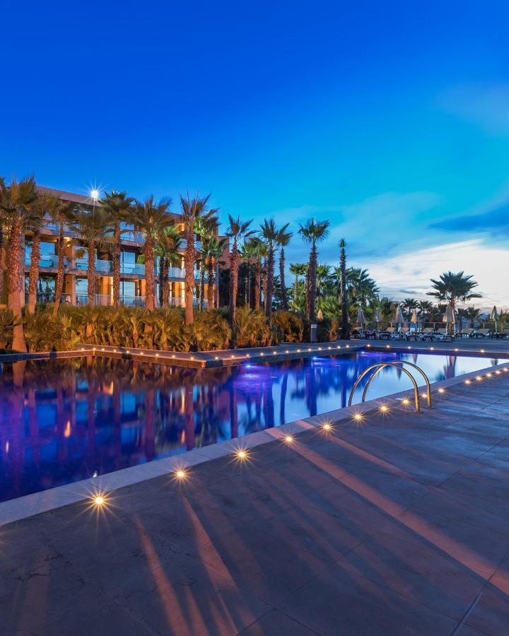 Ap Lago Montargil - Conference & Spa Exterior photo The image shows a serene and inviting outdoor area with a large swimming pool that features a beautiful blue hue, likely illuminated by lights. Surrounding the pool are well-maintained palm trees and lush greenery, creating a tropical atmosphere. In 